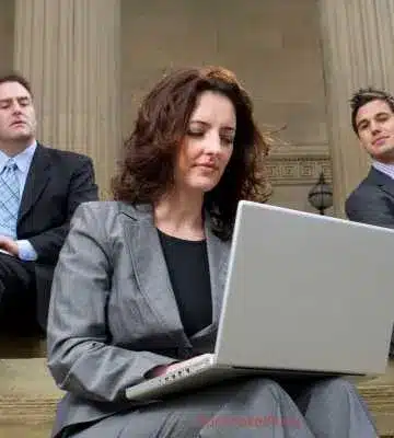 A képen három üzletember látható, akik egy épület lépcsőin ülnek. Középen egy nő van, aki egy laptopon dolgozik, figyelmesen nézve a képernyőt. Mellette két férfi ül, akik közömbös vagy irigykedő tekintettel néznek rá. A háttérben az épület oszlopai láthatók, ami hivatalos vagy üzleti környezetet sugall.