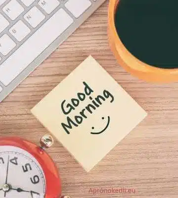 Reggeli köszöntések. A képen egy fa íróasztal látható billentyűzettel, egy piros ébresztőórával és egy narancssárga kávéscsészével. Az asztalon egy jegyzetlap található, amelyen a "Good Morning" (Jó reggelt) felirat szerepel egy mosolygós arc alatt.