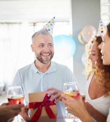 Vicces szülinapi köszöntők férfiaknak. Egy születésnapi partin készült kép, ahol a középpontban egy idősebb férfi áll, aki éppen egy ajándékot kap. A férfi vidáman mosolyog, kezében egy piros szalaggal átkötött ajándékdobozt tart. Körülötte három másik személy látható, akik pezsgőspoharat tartanak a kezükben és szintén mosolyognak. A háttérben lufik és más parti dekorációk díszítik a helyiséget. Mindenki ünnepi party sapkát visel, ami színes és vidám hangulatot kölcsönöz a képnek.