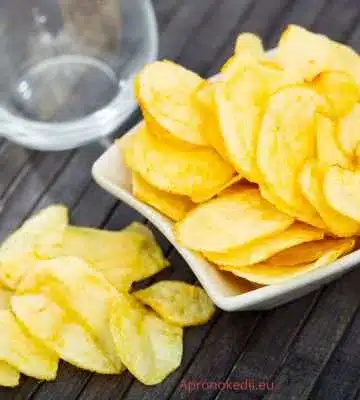 Ez a kép chips-szerűen vékonyra szeletelt burgonyát mutat egy kis tálban. A burgonya szeletek aranysárgák, és egy része a tálból kiborult az asztalra. A háttérben egy üveg látható, amely homályosan van fókuszban.